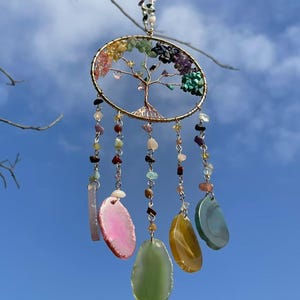 May include: A gold wire tree of life dreamcatcher with colourful gemstone beads and agate slices hanging from it. The dreamcatcher is hanging from a branch against a blue sky.