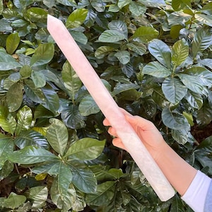 Op de afbeelding: Een grote, roze kristallen toverstaf met een puntige punt. De toverstaf wordt in een hand gehouden tegen een achtergrond van groene bladeren.