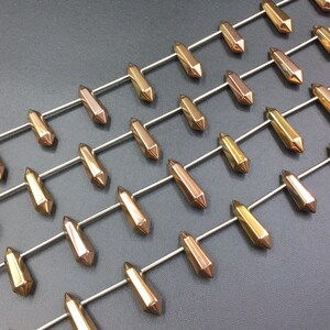 Puede incluir: Primer plano de una hebra de cuentas de cristal doradas y de oro rosa. Las cuentas son facetadas y tienen una forma puntiaguda. Las cuentas están enhebradas en un alambre de plata.