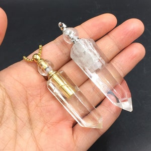 Może przedstawiać: Dwa przezroczyste wisiorek z kryształu kwarcowego z pozłacanymi i srebrnymi nasadkami i łańcuszkami. Wisiorek jest trzymany w dłoni.