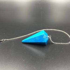 Può includere: Un pendolo di cristallo turchese con una catena d'argento. Il pendolo ha la forma di una punta e ha una piccola perla d'argento nella parte superiore della catena.