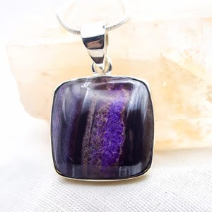 May include: A square pendant with a polished, deep purple stone, set in a silver-colored metal frame. The stone has a unique pattern of lighter purple and white streaks. The pendant hangs from a delicate silver chain, creating a striking piece of jewelry.