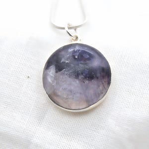 May include: A round pendant necklace featuring a polished, translucent purple and white stone set in a smooth silver-toned metal bezel. The pendant hangs from a simple silver-toned chain, showcasing natural mineral inclusions and varied color depth.