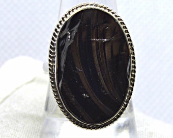 May include: An oval ring featuring a dark brown, striped gemstone set within a twisted metal border. The stone displays a smooth, reflective surface with subtle vertical banding. The word Pixby is visible in the top right corner.