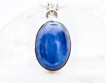 May include: A pendant featuring a smooth, oval-shaped blue kyanite gemstone set in a polished silver frame. The stone displays deep blue hues with subtle internal striations and a reflective surface. A silver bail is attached to the top of the setting.
