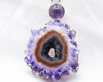 May include: A pendant featuring a raw, purple amethyst geode slice with a dark, hollow center and a brown ring, topped with a smooth, round, polished purple amethyst cabochon set in a silver metal frame with a bail for hanging on a chain.