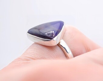 May include: A silver ring featuring a large, triangular, deep purple stone with subtle dark veining. The stone is set in a smooth, polished silver bezel that transitions into a simple, rounded silver band, showcasing a clean and reflective metal finish.