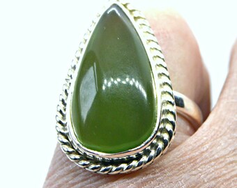 May include: A silver ring featuring a large, smooth, teardrop-shaped olive green stone set in a polished metal bezel. The stone is surrounded by a decorative twisted rope-style silver border, creating a textured frame around the central gem.