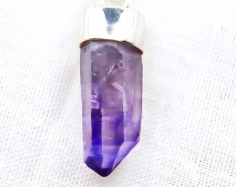 May include: A pendant featuring a raw, elongated amethyst crystal with a deep purple hue that transitions to a lighter shade at the top. The crystal is secured by a polished silver-toned metal cap with a circular bail for attachment to a chain.