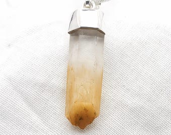 May include: A pendant necklace featuring a long, hexagonal, clear quartz crystal point with a gradient of amber-colored inclusions at the base. The crystal is secured by a polished silver-toned metal cap and hangs from a matching silver-toned chain.