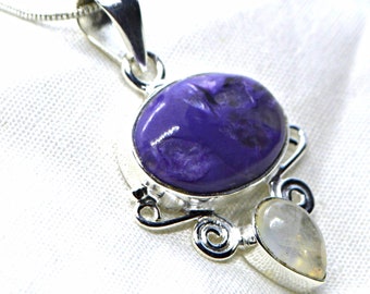 May include: A silver pendant featuring a large oval purple charoite stone set above a smaller pear-shaped iridescent moonstone. The stones are encased in polished silver metal with decorative scrollwork accents and a silver chain bail at the top.