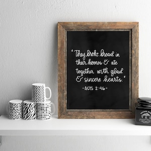 May include: A black framed chalkboard with a white handwritten quote: "They broke bread in their homes & ate together with glad & sincere hearts." -ACTS 2:46-. The chalkboard is on a white shelf with four black and white patterned mugs and a black tea jar.