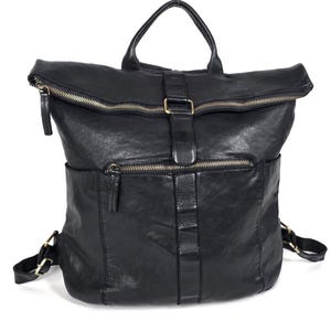 Pode incluir: Uma mochila de couro preta com fecho superior dobrável com zíper, uma alça superior e um bolso frontal com zíper. A bolsa inclui uma tira central vertical com detalhe de fivela e alças de ombro ajustáveis com ferragens metálicas.