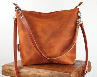 May include: A brown leather shoulder bag with a long strap. The bag is sitting on a wooden stool.