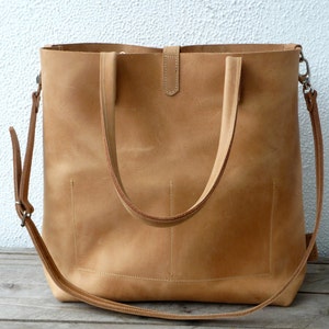 Puede incluir: Un bolso tote de cuero color marr&oacute;n claro con dos asas superiores y una correa de hombro larga ajustable. El bolso tiene una cremallera y un bolsillo frontal.