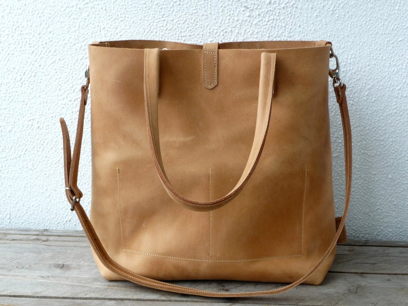 Puede incluir: Un bolso tote de cuero color marr&oacute;n claro con dos asas superiores y una correa de hombro larga ajustable. El bolso tiene una cremallera y un bolsillo frontal.