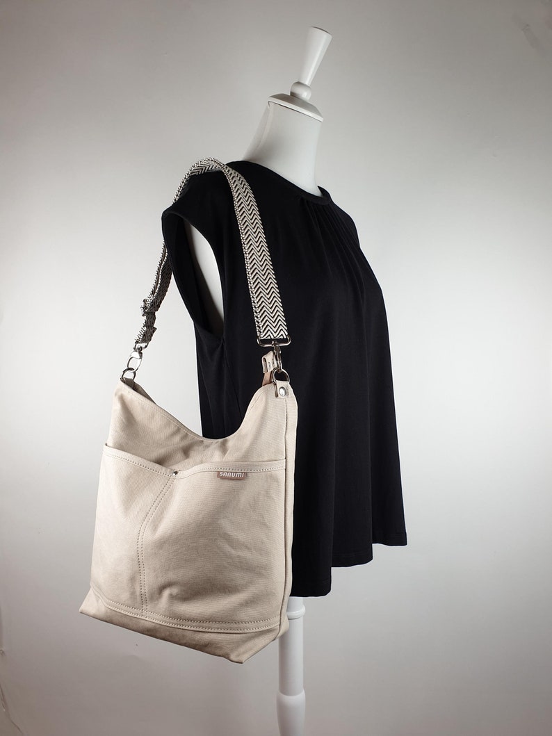 Umhängetasche in beige aus Canvas hängt über der Schulter einer Schneiderpuppe, die ein schwarzes T-shirt anhat.