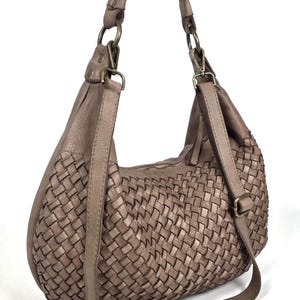 May include: Taupe-colored shoulder bag with a woven design. The bag features a long, adjustable strap and a shorter handle. The bag has a zipper closure and metal hardware. The bag is made of a soft, leather-like material.