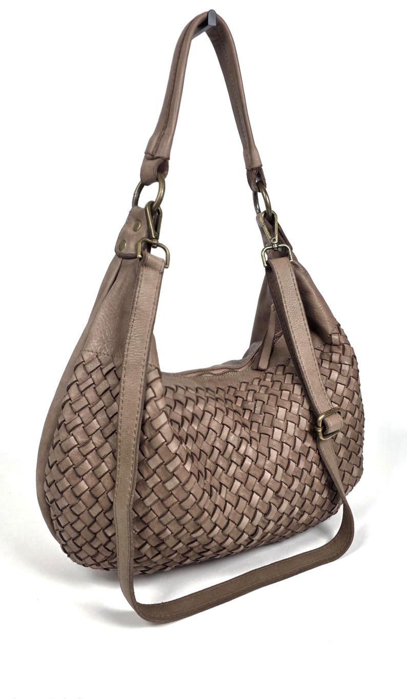 May include: Taupe-colored shoulder bag with a woven design. The bag features a long, adjustable strap and a shorter handle. The bag has a zipper closure and metal hardware. The bag is made of a soft, leather-like material.