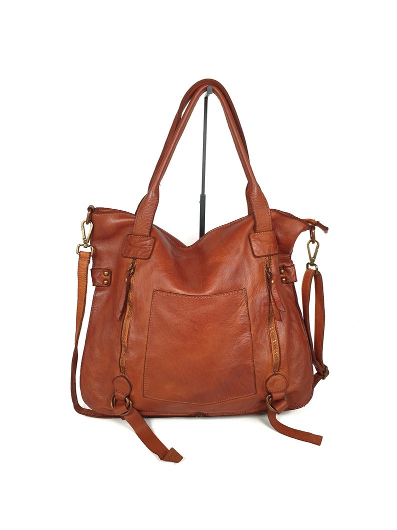 May include: A brown leather tote bag with two handles and a detachable shoulder strap. The bag has two exterior zippered pockets and a large pocket on the back. The bag is made of a distressed leather.