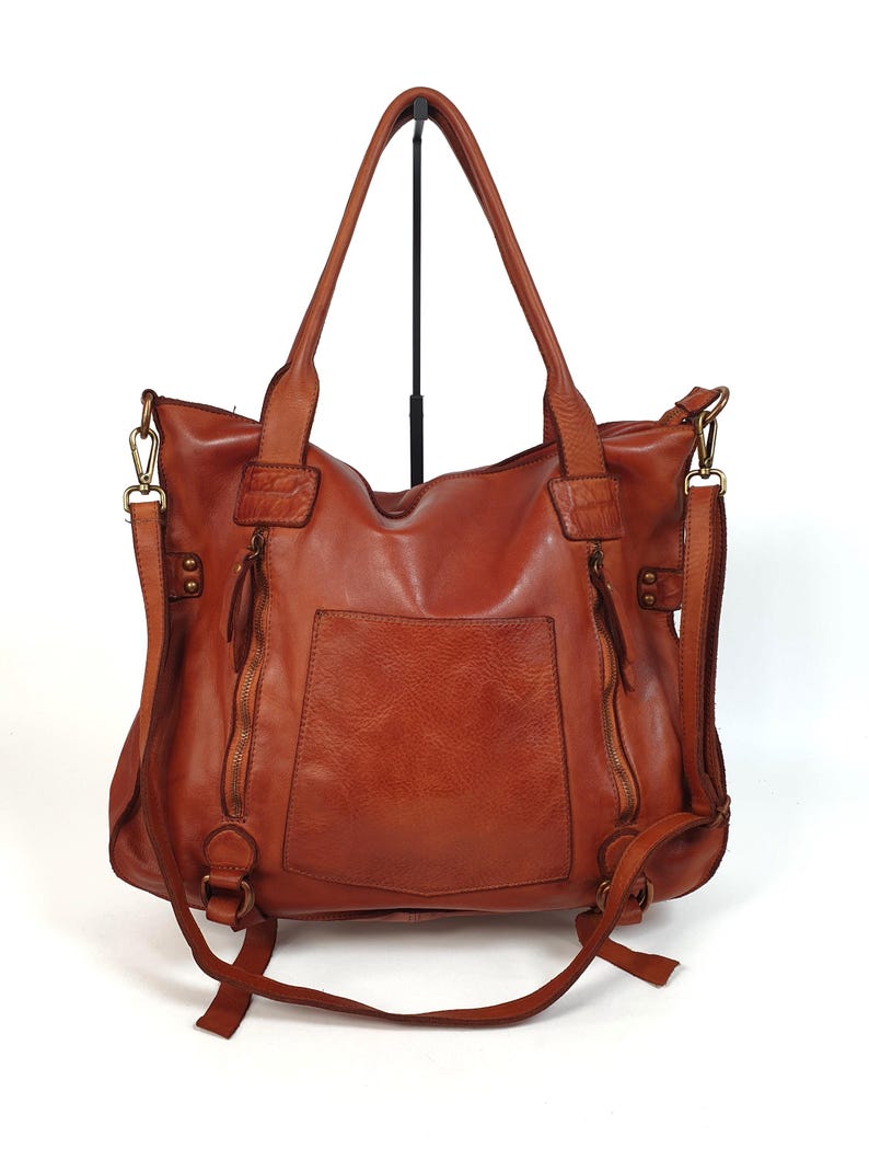 May include: A brown leather tote bag with a front pocket and two zippered compartments. The bag has two top handles and a detachable shoulder strap. The bag is made of a soft, distressed leather.