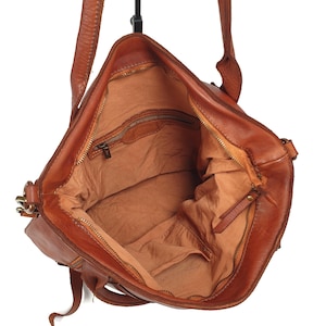 May include: Open view of a brown leather tote bag with a long shoulder strap. The bag has a zipper closure and a fabric lining. The interior reveals a pocket and a spacious compartment. The leather appears worn, adding to its vintage appeal.