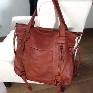 Das Bild zeigt eine große Leder Tote Bag für Damen in cognac braun der Marke sanumileathergoods. Diese Tasche ist 44 cm breit, 37 cm hoch und 13 cm tief und hängt mit beiden Schulterhenkeln an einem Sessel in einem Wohnraum. Diese weiche Ledertasche schließt oben mit einem Reißverschluss. Auf der Vorderseite der Schultertasche befinden sich zwei kleine Reißverschlussfächer und ein Steckfach. Zusätzlich hat die Tasche einen langen Leder Umhängegurt. Die Metallteile sind messingfarben.