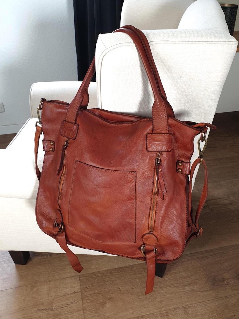 Das Bild zeigt eine große Leder Tote Bag für Damen in cognac braun der Marke sanumileathergoods. Diese Tasche ist 44 cm breit, 37 cm hoch und 13 cm tief und hängt mit beiden Schulterhenkeln an einem Sessel in einem Wohnraum. Diese weiche Ledertasche schließt oben mit einem Reißverschluss. Auf der Vorderseite der Schultertasche befinden sich zwei kleine Reißverschlussfächer und ein Steckfach. Zusätzlich hat die Tasche einen langen Leder Umhängegurt. Die Metallteile sind messingfarben.