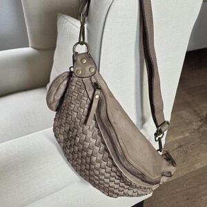 Puede incluir: Ri&ntilde;onera de cuero tejido en color topo neutro. La bolsa presenta una forma curva, un cierre de cremallera y una correa ajustable. La bolsa est&aacute; colgada en el respaldo de un sill&oacute;n blanco.