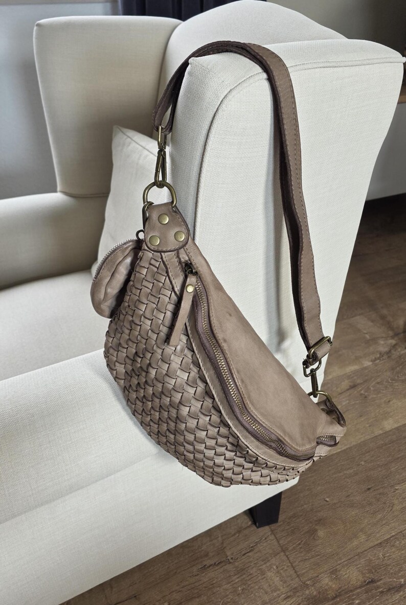 Puede incluir: Ri&ntilde;onera de cuero tejido en color topo neutro. La bolsa presenta una forma curva, un cierre de cremallera y una correa ajustable. La bolsa est&aacute; colgada en el respaldo de un sill&oacute;n blanco.