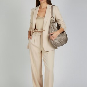 May include: A woman wearing a beige pantsuit with a matching blazer and a satin top. She is carrying a woven gray handbag. The outfit is completed with white sneakers.