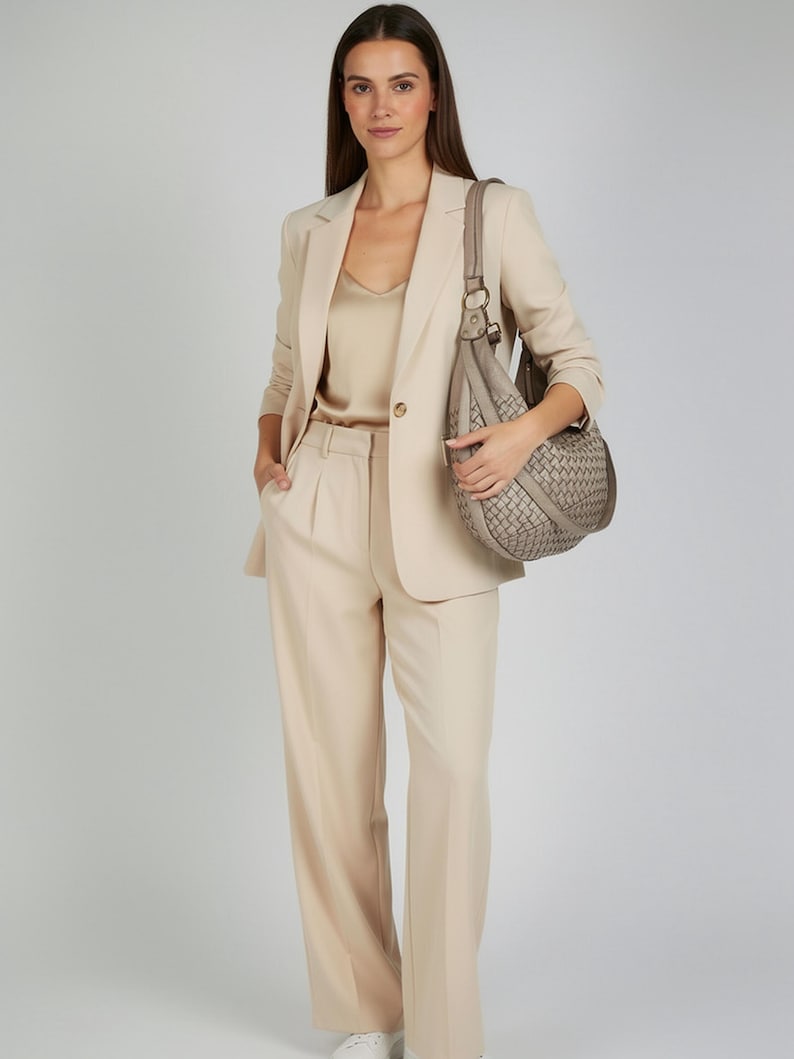 May include: A woman wearing a beige pantsuit with a matching blazer and a satin top. She is carrying a woven gray handbag. The outfit is completed with white sneakers.