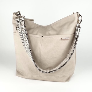 Eine mittelgroße Canvas Crossbodytasche in der Farbe greige für Damen mit einem schwarz-weiß gemusterten Zickzack-Gurt steht vor weißem Hintergrund. Die Tasche ist aus festem Segeltuchstoff, ist sehr robust und steht von selbst. Die Umhängetasche hat 2 Einsteckfächer auf der Vorderseite und ein kleines Etikett mit mit dem Markennamen "SANUMI". Der Gurt ist mit silberfarbenen Metall-Karabinerhaken befestigt.
