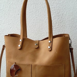 Puede incluir: Un bolso tote de cuero marr&oacute;n claro con una correa larga y un bolsillo m&aacute;s peque&ntilde;o en la parte delantera. El bolso tiene herrajes plateados y un par de gafas de sol cuelgan del bolsillo.