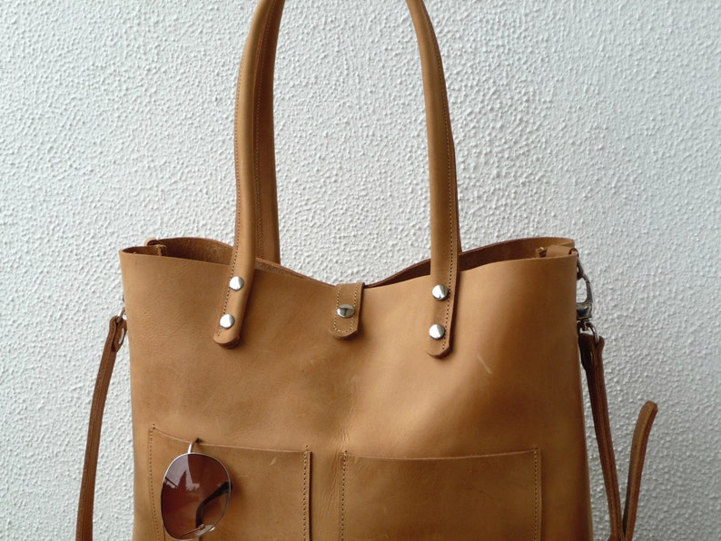 Puede incluir: Un bolso tote de cuero marr&oacute;n claro con una correa larga y un bolsillo m&aacute;s peque&ntilde;o en la parte delantera. El bolso tiene herrajes plateados y un par de gafas de sol cuelgan del bolsillo.