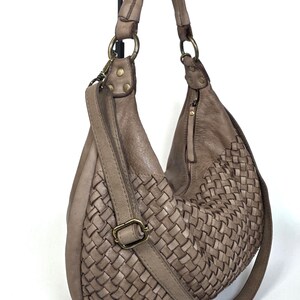 May include: A taupe-colored leather shoulder bag with a woven design on the front. The bag has a long adjustable strap, a top handle, and brass-colored hardware. The bag is hanging against a white background.