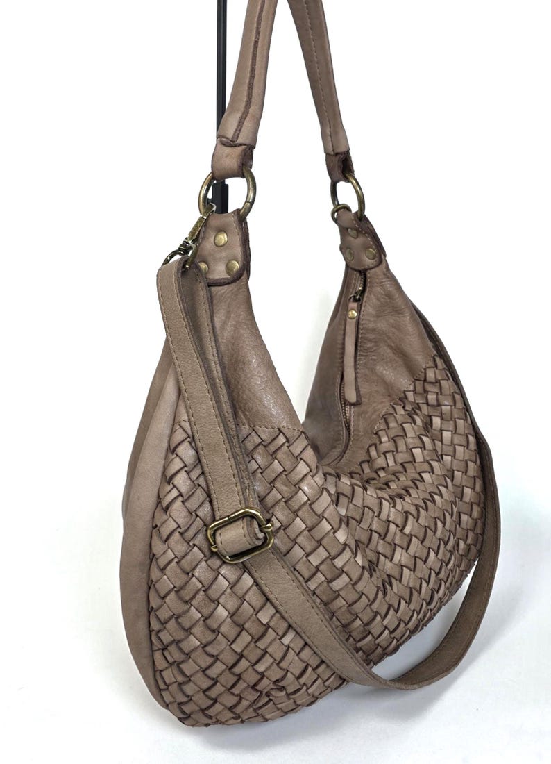 May include: A taupe-colored leather shoulder bag with a woven design on the front. The bag has a long adjustable strap, a top handle, and brass-colored hardware. The bag is hanging against a white background.