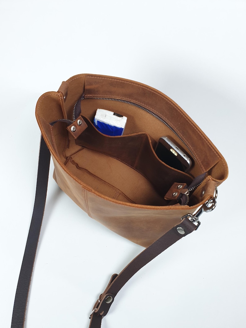Op de afbeelding: Bruine leren schoudertas met een donkerbruine riem en zilverkleurige hardware. De tas is open en onthult binnenvakken en een ritssluiting. Een smartphone en een kleine verpakking zijn zichtbaar.