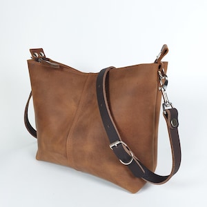 Op de afbeelding: Bruine leren crossbodytas met een verstelbare donkerbruine riem en zilverkleurige hardware. De tas heeft een rechthoekige vorm met een ritssluiting en een gestikte naad aan de voorkant.