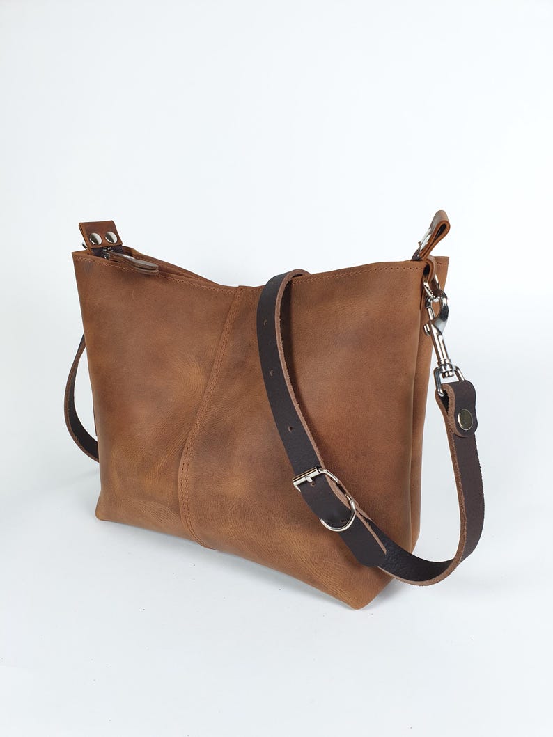 Op de afbeelding: Bruine leren crossbodytas met een verstelbare donkerbruine riem en zilverkleurige hardware. De tas heeft een rechthoekige vorm met een ritssluiting en een gestikte naad aan de voorkant.