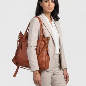 May include: A woman wearing a tan suit and carrying a brown leather tote bag and a matching shoulder bag. The tote bag has multiple zippers and a long strap. The shoulder bag has a long adjustable strap. The background is plain white.