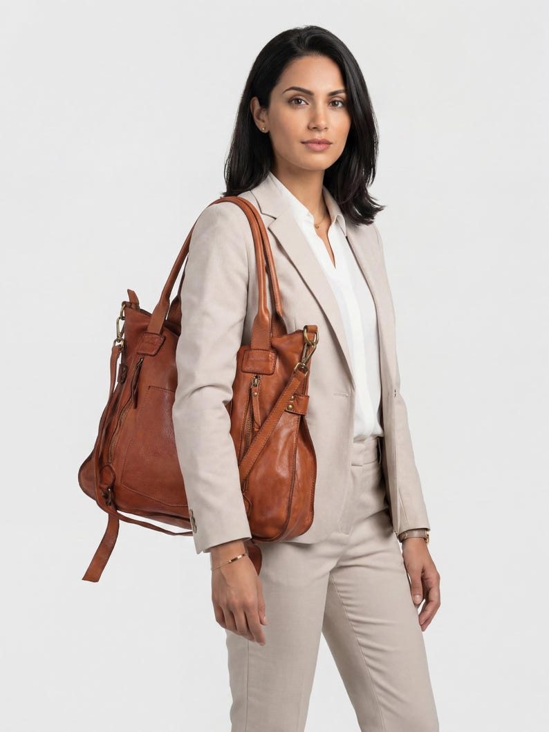May include: A woman wearing a tan suit and carrying a brown leather tote bag and a matching shoulder bag. The tote bag has multiple zippers and a long strap. The shoulder bag has a long adjustable strap. The background is plain white.
