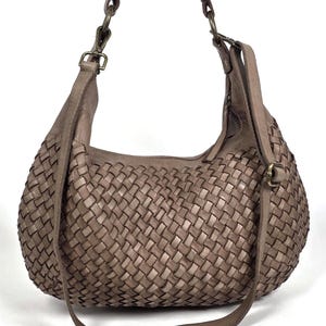 May include: A woven leather shoulder bag in a neutral taupe color. The bag features a large, rounded shape with a woven pattern across the body. It has a long, adjustable strap and a shorter shoulder strap, both in matching leather.