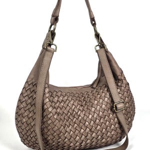 May include: A taupe-colored woven leather shoulder bag with a curved shape. The bag features a long, adjustable strap and a woven pattern across the body. The hardware is a bronze color.