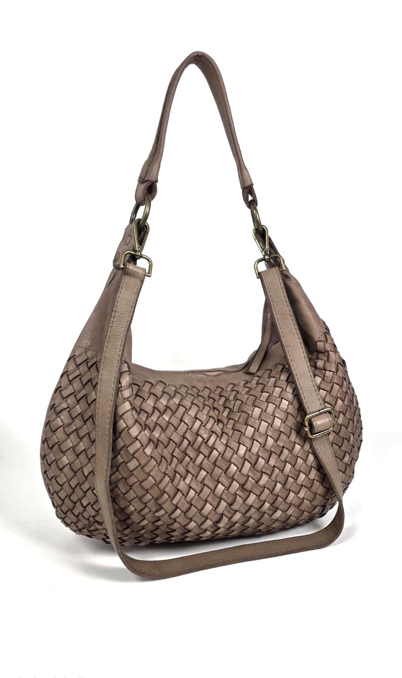 May include: A taupe-colored woven leather shoulder bag with a curved shape. The bag features a long, adjustable strap and a woven pattern across the body. The hardware is a bronze color.