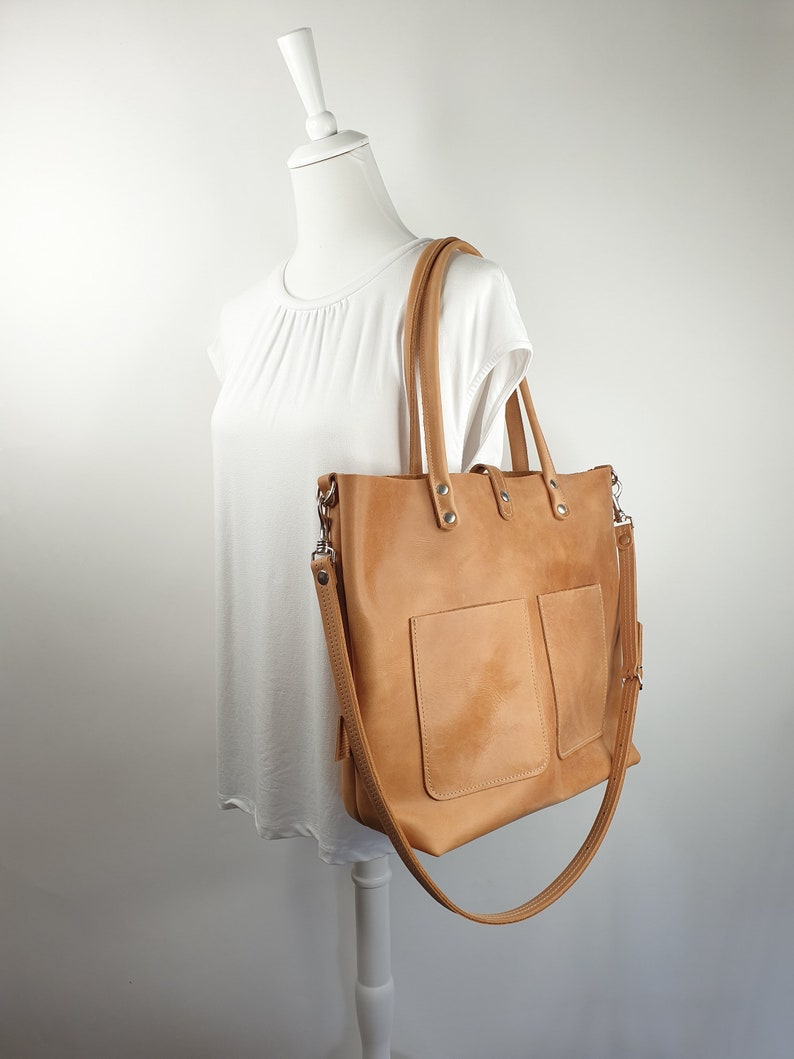 Puede incluir: Un bolso tote de cuero marr&oacute;n claro con una correa larga y un bolsillo frontal. El bolso est&aacute; en un maniqu&iacute; blanco.