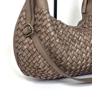 May include: A woven leather shoulder bag in a neutral taupe color. The bag features a braided design on the main body and a long strap. A gold-colored buckle secures the strap. The bag is a stylish accessory.