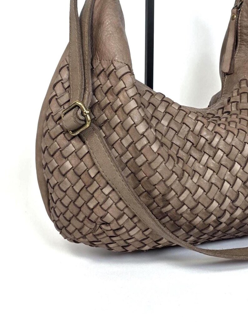 May include: A woven leather shoulder bag in a neutral taupe color. The bag features a braided design on the main body and a long strap. A gold-colored buckle secures the strap. The bag is a stylish accessory.