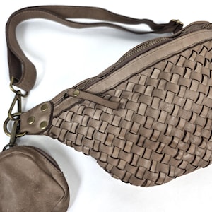 Puede incluir: Ri&ntilde;onera de cuero tejido en color topo neutro. La bolsa cuenta con un compartimento principal con cremallera, una correa ajustable y un peque&ntilde;o monedero adjunto. El dise&ntilde;o de la bolsa incluye un patr&oacute;n tejido y herrajes de color lat&oacute;n.