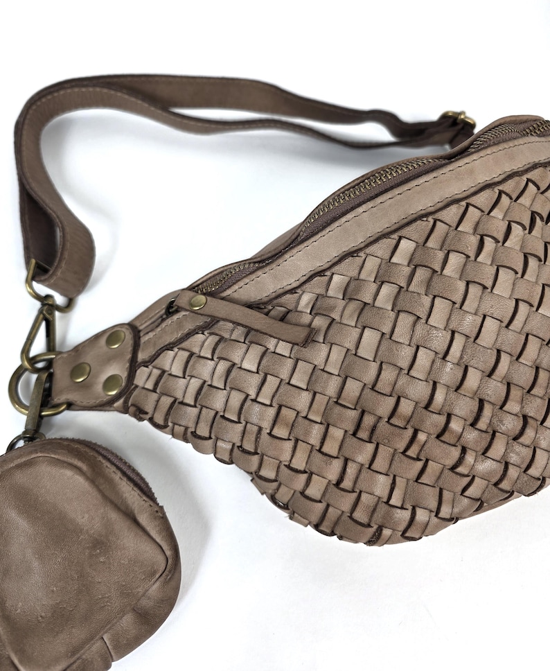 Puede incluir: Ri&ntilde;onera de cuero tejido en color topo neutro. La bolsa cuenta con un compartimento principal con cremallera, una correa ajustable y un peque&ntilde;o monedero adjunto. El dise&ntilde;o de la bolsa incluye un patr&oacute;n tejido y herrajes de color lat&oacute;n.